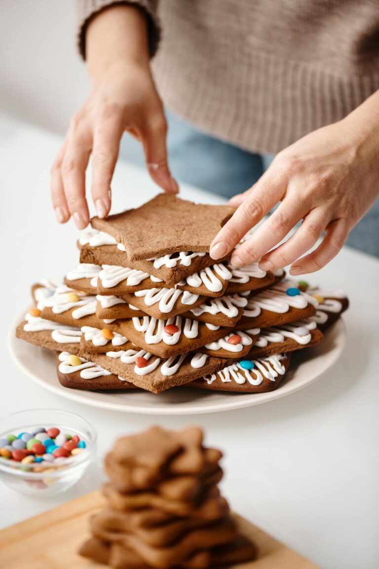 Hands decorating a stack of gingerbread cookies with icing and candy for Christmas festivities.